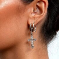 Model ear wearing silver-toned hoop earrings including ornate filigree cross charm drop, close detail