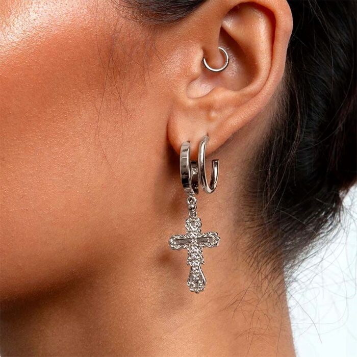 Model ear wearing silver-toned hoop earrings including ornate filigree cross charm drop, close detail