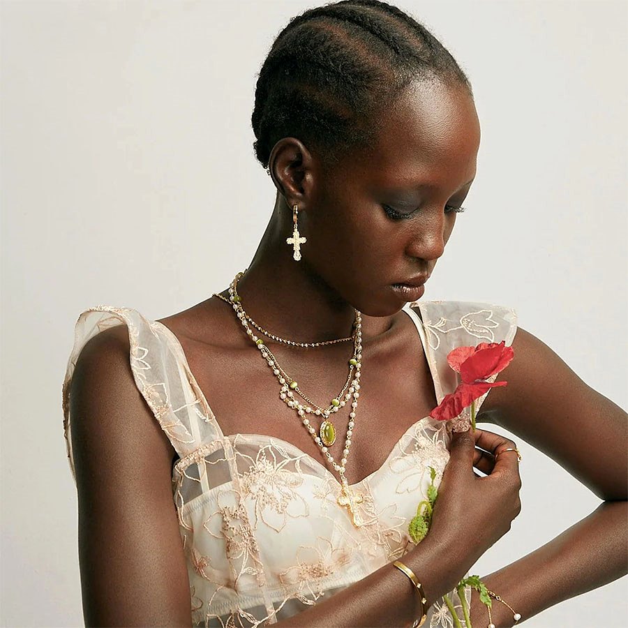 Model wearing Everette Cross Necklace in Gold with layered pearl necklaces and a gold cross earring, holding a red poppy