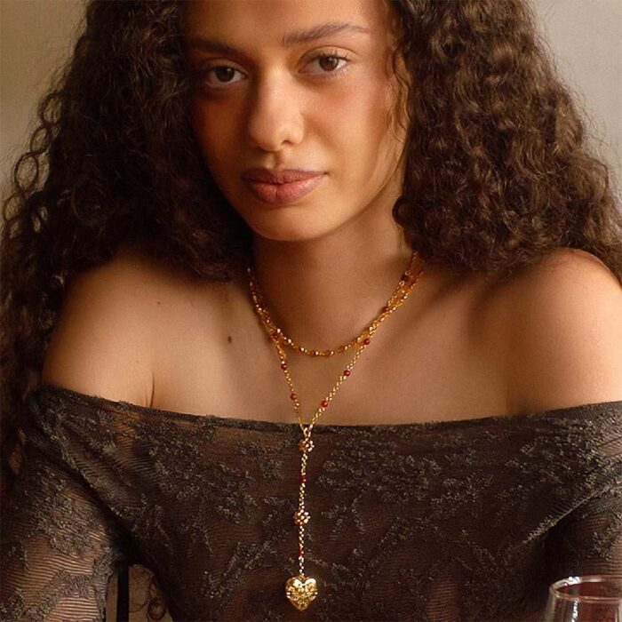 Model wearing a layered gold necklace with red beads and a heart pendant