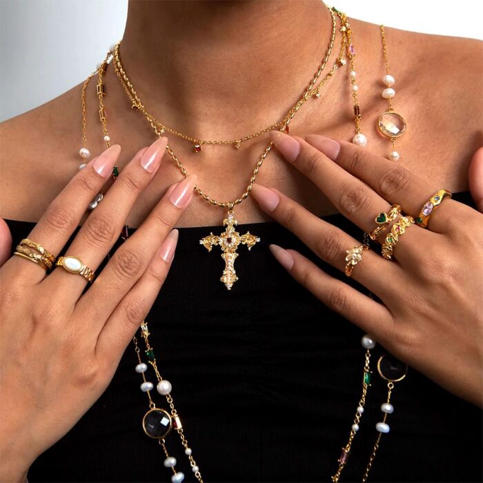Model wearing the Aphrodite Cross Necklace, a gold-toned ornate cross pendant layered with pearl and gemstone necklaces and stacked rings.